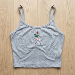 May include: A gray crop top with thin straps. The top features an embroidered design of a pig with a rooster on its head. The design is in shades of blue, red, yellow, and white. The top is made of a soft, comfortable material.