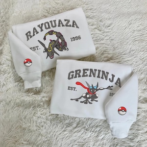 May include: Two white sweatshirts with embroidered designs. One features "RAYQUAZA" and a dragon graphic, the other "GRENINJA" with a ninja-like character. Both have "EST. 1996" and a Pokeball on the sleeve.