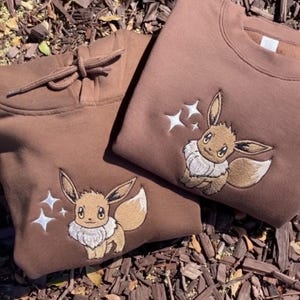 Op de afbeelding: Twee bruine sweatshirts, een hoodie en een crewneck, beide met een geborduurd Eevee-ontwerp. De Eevee heeft bruin haar, een witte kraag en een pluizige staart. Witte steraccenten zijn boven het karakter geborduurd.