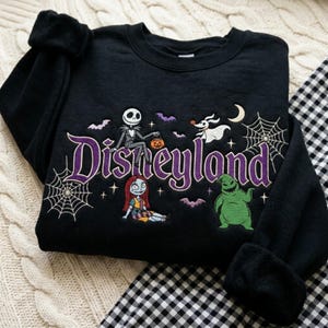 May include: A black crewneck sweatshirt featuring an embroidered design with the purple text Disneyland. The graphic includes Jack Skellington, Sally, Zero, and Oogie Boogie from The Nightmare Before Christmas, along with spiderwebs and bats.
