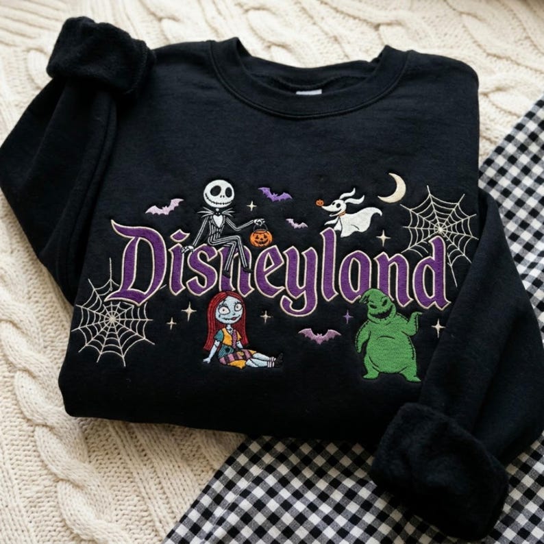 May include: A black crewneck sweatshirt featuring an embroidered design with the purple text Disneyland. The graphic includes Jack Skellington, Sally, Zero, and Oogie Boogie from The Nightmare Before Christmas, along with spiderwebs and bats.