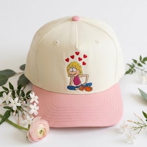 May include: Cream-colored baseball cap with a pink brim and button. The cap features an embroidered cartoon girl with hearts above her head. The design is set against a white background with floral accents.