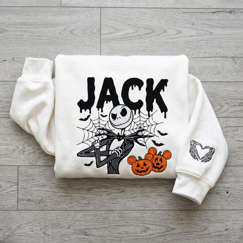 May include: A white long-sleeve sweatshirt featuring a black graphic of Jack Skellington with a spiderweb background and the text JACK in a dripping font. Two orange Mickey Mouse-shaped pumpkins are below, and a skeleton hand heart is on the sleeve.