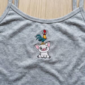 May include: A gray tank top with thin straps. The tank top features an embroidered design of a cartoon pig with a rooster on its head. The rooster has blue, yellow, and red coloring. The pig is white with pink ears and a smiling face.