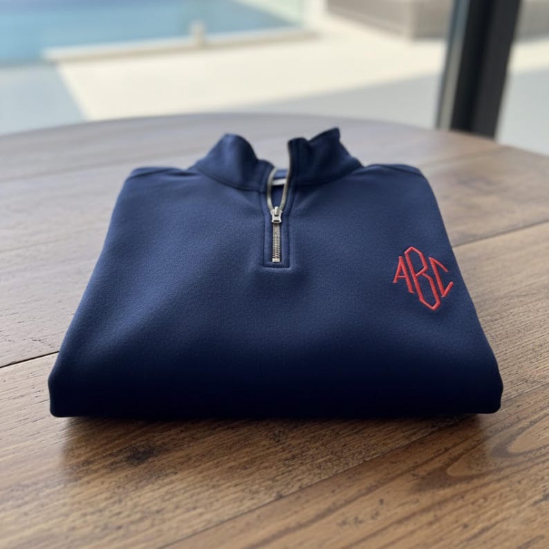 May include: Navy blue quarter-zip pullover with a red embroidered monogram on the chest. The folded sweater has a high collar and a silver zipper. The garment is made of a soft, textured fabric, perfect for casual wear.