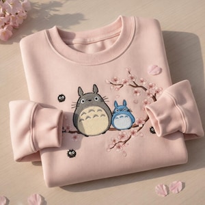 May include: A pale pink sweatshirt with an embroidered design of two cartoon characters, one grey and one blue, with a floral branch. The sweatshirt has a crew neck and long sleeves with ribbed cuffs. Pink flower petals are scattered around.