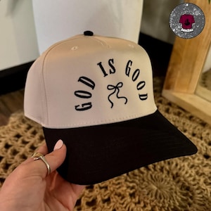 May include: A beige and black baseball cap with the words "GOD IS GOOD" embroidered in black. The cap has a black brim and a small black button on top. The cap is being held by a person.