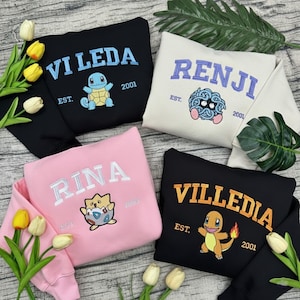 May include: Four sweatshirts in black, cream, pink, and black. Each features a name in arched lettering, a cartoon character, and the year 2001 or 2003. The black sweatshirt has "VI LEDA" and a blue turtle. The cream sweatshirt has "RENJI" and a blue creature.