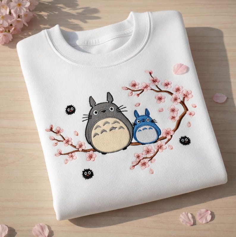 May include: White sweatshirt with an embroidered design of two Totoro characters and a flowering cherry blossom branch. The larger Totoro is grey with a beige belly, and the smaller one is blue. Black soot sprites are also embroidered.