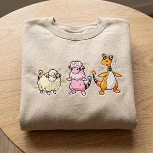 May include: Beige crewneck sweatshirt featuring embroidered cartoon sheep characters. The design includes a yellow sheep, a pink sheep, and an orange and white sheep-like creature. The sweatshirt is folded on a wooden surface.