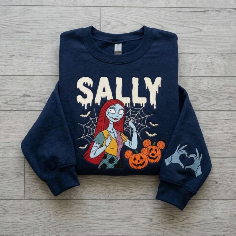 May include: A navy blue long-sleeve sweatshirt featuring the text SALLY in a white dripping font. The graphic shows a character with long red hair, a patchwork dress, and a spiderweb background, alongside two orange jack-o'-lanterns and hand silhouettes.