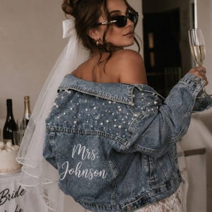 Puede incluir: Una chaqueta de mezclilla azul con adornos de perlas blancas en la espalda y los hombros. El texto Mrs. Johnson está impreso en letra cursiva blanca en la parte inferior de la espalda. Un velo blanco con un lazo está sujeto a la chaqueta, que se lleva sobre una prenda de encaje blanco.