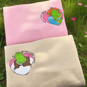 May include: Two sweatshirts, one pink and one beige, embroidered with cartoon characters inside a heart shape. The pink sweatshirt has a green character with orange hair. The beige sweatshirt has a green character wearing a hat. Text in Portuguese is also included.