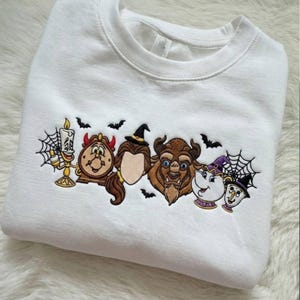 Puede incluir: Una sudadera blanca de cuello redondo con un diseño bordado de personajes animados que incluye una vela, un reloj con cuernos rojos, una persona de cabello castaño con sombrero de bruja, una bestia, una tetera y una taza, con telarañas y murciélagos negros.