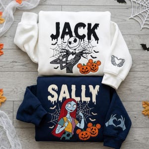 May include: A white sweatshirt featuring Jack Skellington, spiderwebs, bats, and orange pumpkins with the text JACK, plus a sleeve print of skeleton hands forming a heart. A navy blue sweatshirt featuring Sally, spiderwebs, bats, and orange pumpkins with the text SALLY, plus a sleeve print of stitched hands forming a heart.