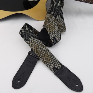 Puede incluir: Una correa de guitarra negra y gris con un patrón de piel de serpiente. La correa tiene extremos de cuero negro con un solo agujero en cada extremo.