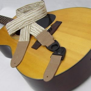 Puede incluir: Una correa de guitarra beige y marrón con un patrón a rayas y un cierre de correa de plástico negro. La correa está unida a una guitarra acústica.