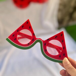 May include: Novelty sunglasses in the shape of watermelon slices. The frames are red with black seed details and green edges, with red lenses. A playful accessory for summer or themed parties.
