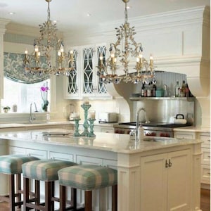 Puede incluir: Una isla de cocina blanca con una encimera blanca y un armario blanco con dos puertas. Tres taburetes de bar de madera con un asiento de tela a cuadros azul y blanco están frente a la isla. Dos lámparas de araña de cristal cuelgan sobre la isla.