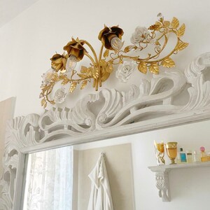 May include: A gold metal wall sconce with a floral design featuring roses and leaves. The sconce has five light bulbs and is mounted on a white wall.