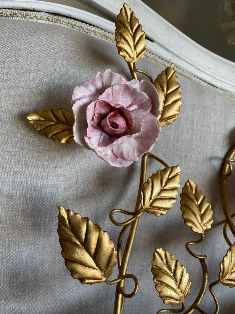 Puede incluir: Una decoraci&oacute;n de pared de metal dorado con una rosa de porcelana rosa y hojas doradas.