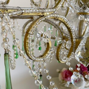 May include: A chandelier featuring a gold metal frame with scrolling arms, adorned with clear glass beads, flower-shaped glass accents, and long, tapered emerald green glass drops. The structure is held together by thin wire connectors.