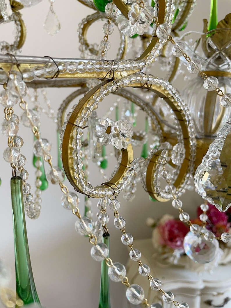 May include: A chandelier featuring a gold metal frame with scrolling arms, adorned with clear glass beads, flower-shaped glass accents, and long, tapered emerald green glass drops. The structure is held together by thin wire connectors.