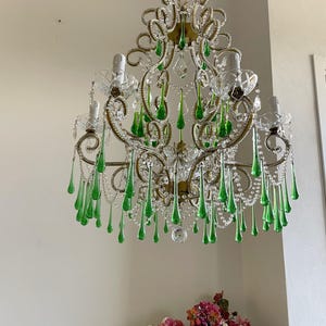 May include: A multi-armed chandelier featuring ornate gold-toned metal scrollwork, clear glass bobeches, and numerous dangling emerald green teardrop-shaped glass crystals. The fixture is suspended by a metal chain and includes clear glass beads.