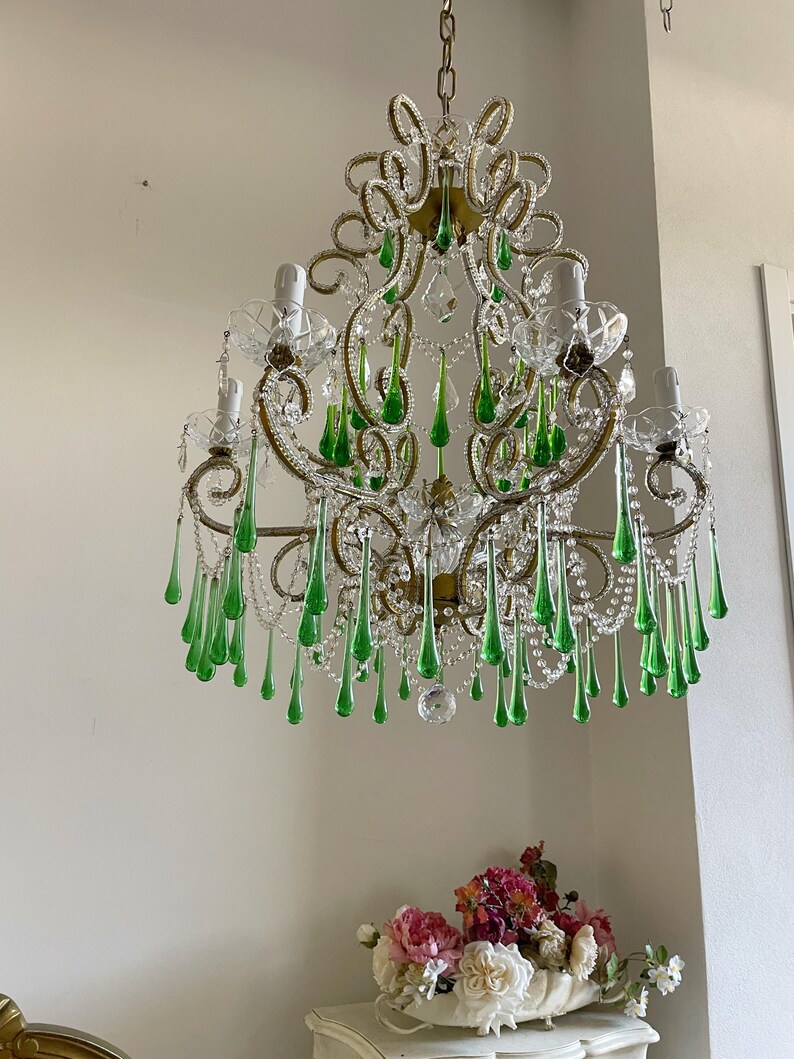 May include: A multi-armed chandelier featuring ornate gold-toned metal scrollwork, clear glass bobeches, and numerous dangling emerald green teardrop-shaped glass crystals. The fixture is suspended by a metal chain and includes clear glass beads.