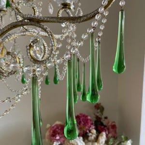 May include: A chandelier featuring curved gold-toned metal arms adorned with clear glass bead strands and multiple elongated, teardrop-shaped green glass pendants. The fixture includes white candle-style light sockets and clear glass accents.