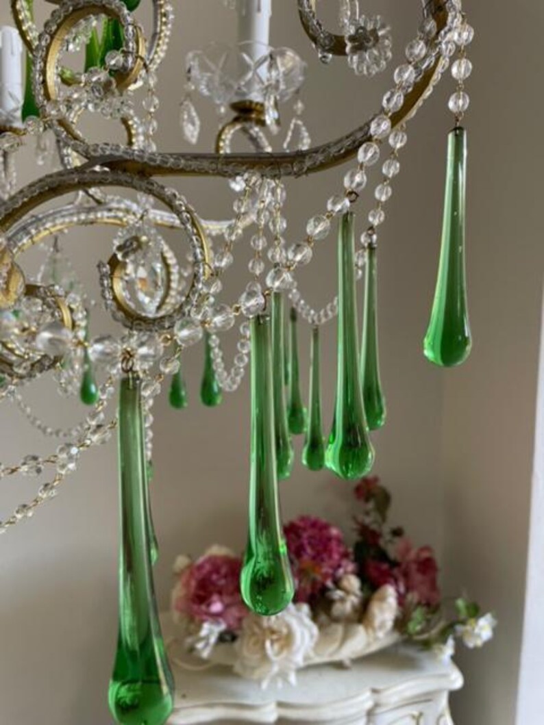 May include: A chandelier featuring curved gold-toned metal arms adorned with clear glass bead strands and multiple elongated, teardrop-shaped green glass pendants. The fixture includes white candle-style light sockets and clear glass accents.