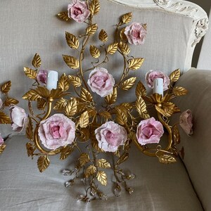 Puede incluir: Una l&aacute;mpara de pared de metal dorado con rosas de porcelana rosa y detalles de cristal. La l&aacute;mpara tiene un dise&ntilde;o floral con hojas y enredaderas.
