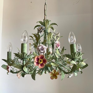 May include: A metal chandelier featuring a sage green finish with sculpted leaves and vines. The fixture is adorned with various ceramic flowers in shades of pink, yellow, and white, and includes four upright candle-style light bulb sockets.