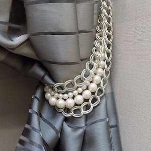 May include: A silver chain curtain tieback with a row of white faux pearls. The tieback is holding back a gray striped curtain.