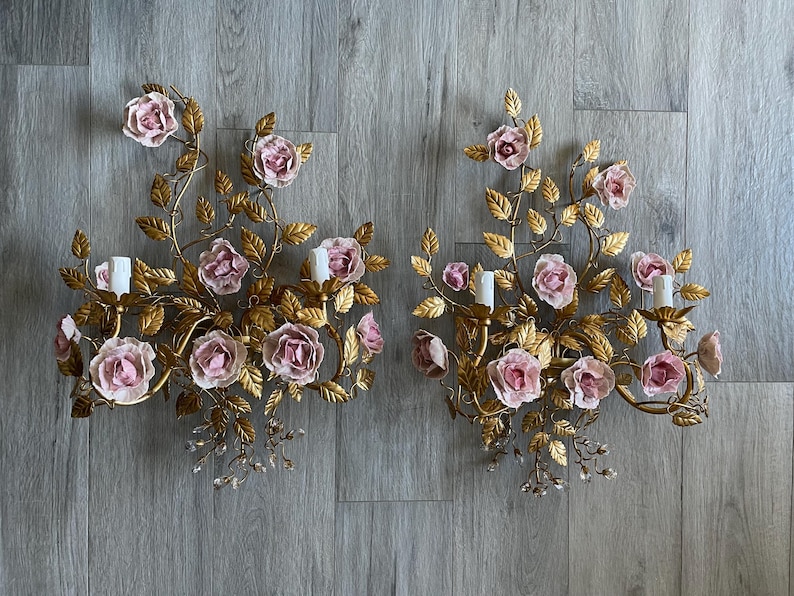 Puede incluir: Un par de apliques de pared doradas con rosas de porcelana rosa y detalles de cristal. Las apliques tienen forma de enredadera floral con hojas y flores.