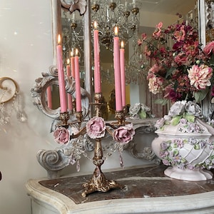 May include: A gold-toned metal candelabra featuring five arms, each adorned with a ceramic pink rose at the base. The candelabra holds five lit pink taper candles and is decorated with clear hanging crystal prisms. The base is ornate and metallic.
