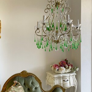 May include: A multi-armed chandelier featuring ornate gold-toned metal scrollwork, clear glass bobeches, and numerous dangling teardrop-shaped green glass crystals. The fixture is suspended by a chain and includes several light bulb sockets.