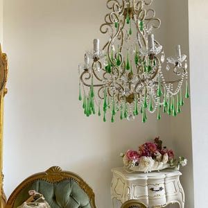 May include: A multi-armed chandelier featuring gold-toned metal scrollwork, clear glass beads, and numerous hanging teardrop-shaped green glass crystals. The fixture includes several upright candle-style light sockets and a central chain suspension.