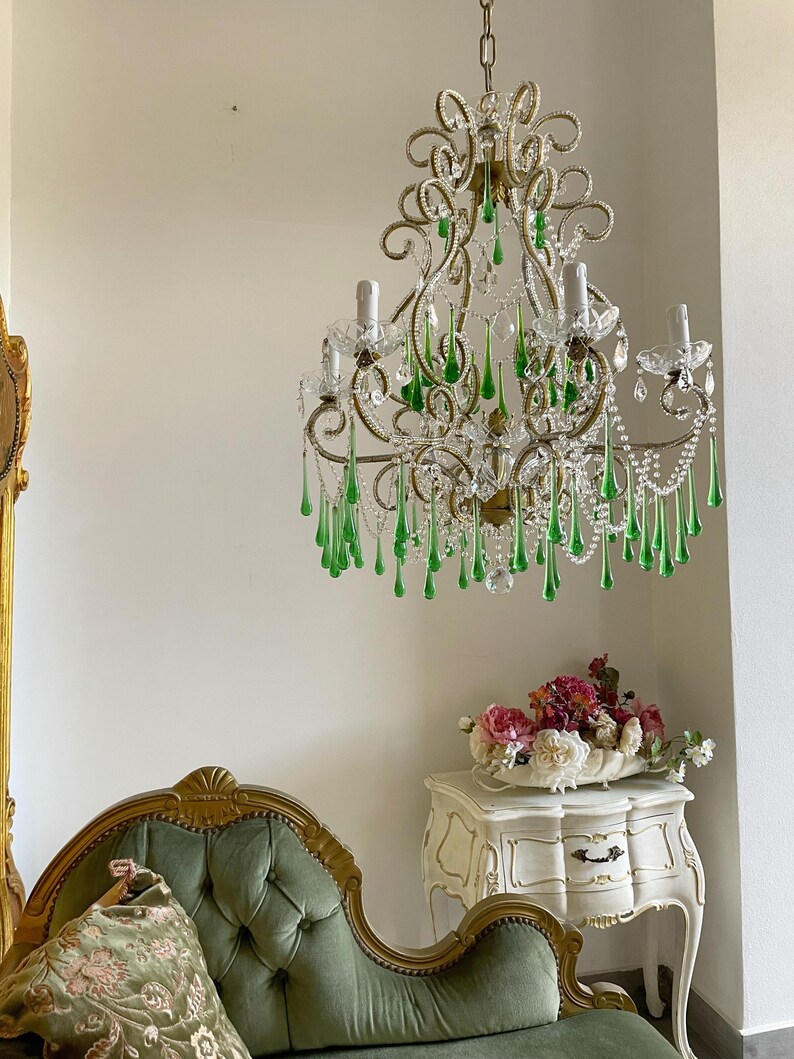May include: A multi-armed chandelier featuring gold-toned metal scrollwork, clear glass beads, and numerous hanging teardrop-shaped green glass crystals. The fixture includes several upright candle-style light sockets and a central chain suspension.