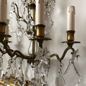 Peut inclure: Une applique murale vintage en laiton et cristal avec quatre bougeoirs et des d&eacute;tails floraux complexes. L'applique est orn&eacute;e de gouttes de cristal claires et d'un pendentif en cristal en forme d'&eacute;toile.