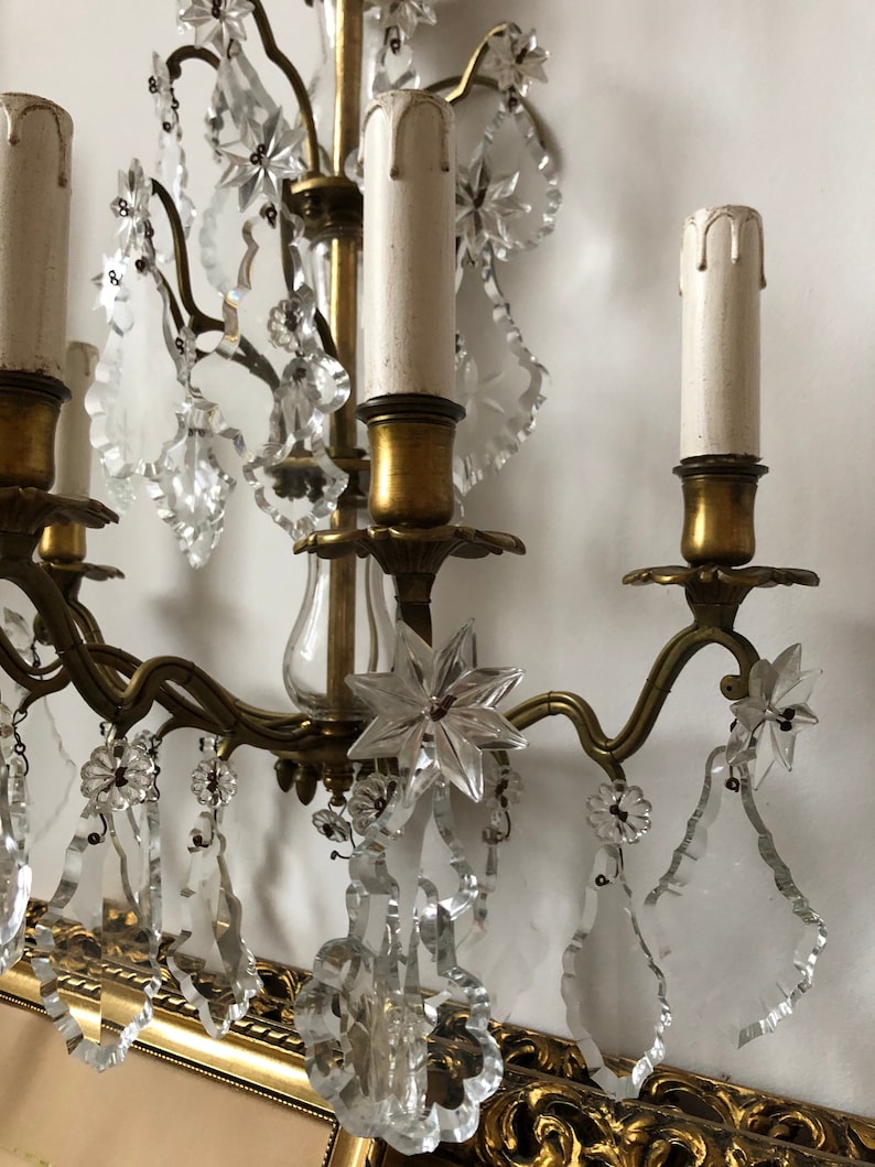 Peut inclure: Une applique murale vintage en laiton et cristal avec quatre bougeoirs et des d&eacute;tails floraux complexes. L'applique est orn&eacute;e de gouttes de cristal claires et d'un pendentif en cristal en forme d'&eacute;toile.