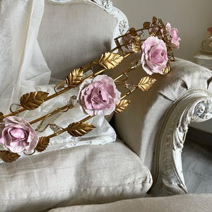 Könnte beinhalten: Eine goldene Wanddekoration aus Metall mit rosafarbenen Porzellanrosen und goldenen Blättern. Die Dekoration wird auf einem hellbeigen Sessel ausgestellt.