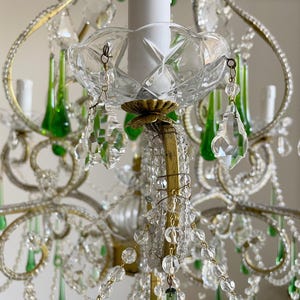 May include: A multi-tiered chandelier featuring a brass frame adorned with clear crystal beads and teardrop-shaped green glass pendants. The fixture includes white candle-style light covers and faceted glass bobeches, creating a layered design.