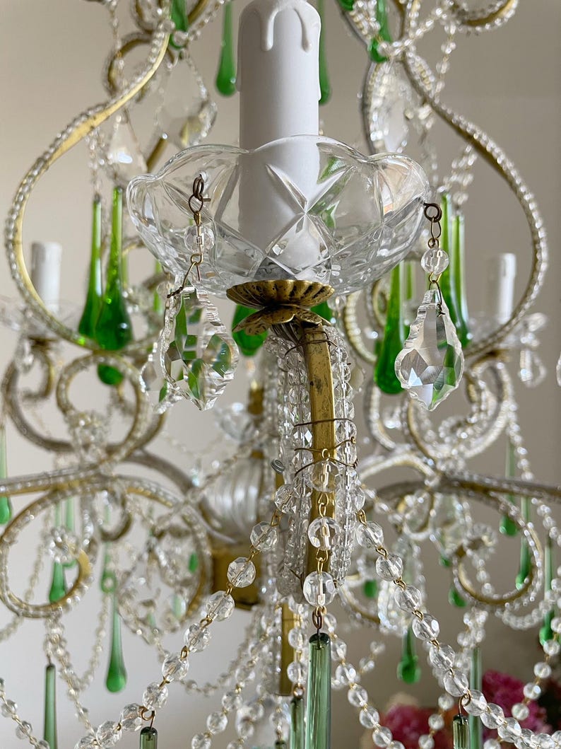 May include: A multi-tiered chandelier featuring a brass frame adorned with clear crystal beads and teardrop-shaped green glass pendants. The fixture includes white candle-style light covers and faceted glass bobeches, creating a layered design.