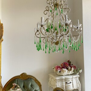 May include: A multi-armed chandelier featuring gold-toned metal scrollwork, clear glass beads, and numerous hanging teardrop-shaped green glass crystals. The fixture includes several upright candle-style light sockets and a central chain suspension.