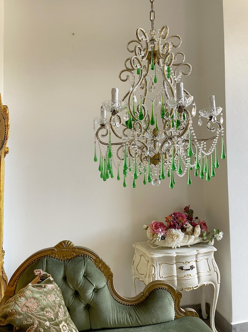 May include: A multi-armed chandelier featuring gold-toned metal scrollwork, clear glass beads, and numerous hanging teardrop-shaped green glass crystals. The fixture includes several upright candle-style light sockets and a central chain suspension.
