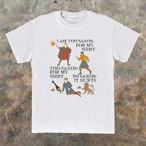 May include: A white short-sleeved t-shirt featuring a graphic print of three medieval-style figures, a sun, a lute, and a lion. The text reads: I AM TOO SAXON FOR MY SHIRT, TOO SAXON FOR MY SHIRT, SO SAXON IT HURTS, arranged around the illustrations.