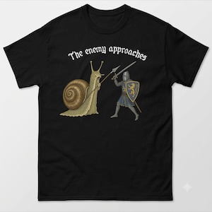 May include: A black short-sleeved t-shirt featuring a graphic of a large snail holding a spear and a knight in armour holding a sword and a blue shield with a gold lion emblem. The text "The enemy approaches" is printed in white above the graphic.