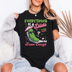 May include: A black t-shirt featuring a graphic of a green pickle wearing sunglasses, lounging on a beach chair under a pink umbrella, holding a cocktail. The text reads: EVERYTHING IS A Dildo IF YOU'RE Brave Enough and EXTRA MOIST.