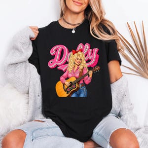 May include: A black short-sleeved t-shirt featuring a graphic illustration of a blonde woman with a red hair bow playing an acoustic guitar. The text Dolly is printed in large pink letters above the illustration. The shirt is worn with a grey cardigan.